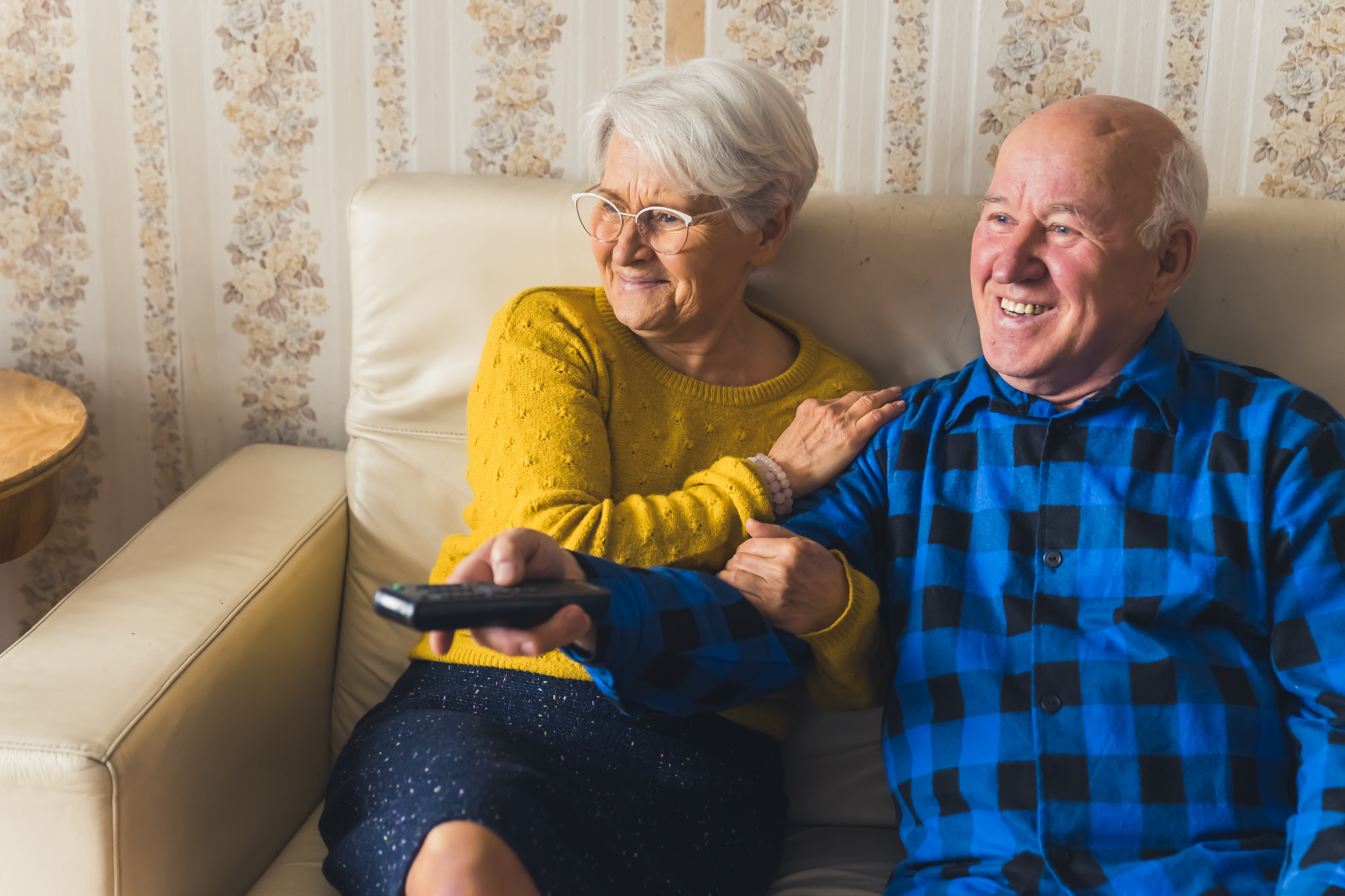 Laughing aged caucasian couple, man and woman a comedy show on watching tv while sitting on a beige leather sofa. Cosy, old-fashioned interior. High quality photo